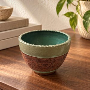 May include: A ceramic bowl featuring a deep teal interior and a textured, sage green upper exterior band. The lower portion of the bowl has a dark, reddish-brown finish with a rough, tactile surface. The rim is detailed with a subtle, rope-like pattern.