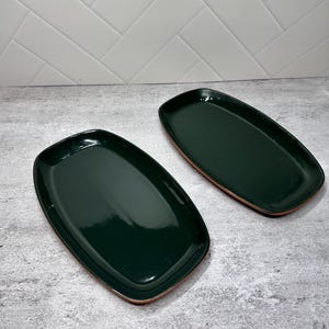 May include: Two rectangular serving platters with rounded corners, featuring a glossy dark forest green interior finish and a contrasting terracotta-colored clay rim along the outer edge, presented as a pair of matching ceramic dishes.