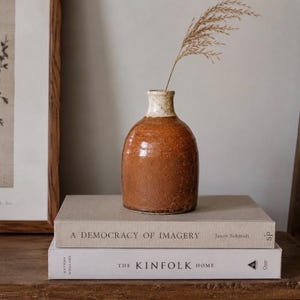 May include: A small, round, rust-colored ceramic vase with a speckled cream-colored neck, holding a single dried grass stem. The vase rests on a stack of two hardcover books titled A Democracy of Imagery and The Kinfolk Home.