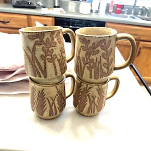May include: Four stacked ceramic mugs with a beige glaze and brown botanical designs. Each mug has a handle and a slightly textured surface. The mugs are arranged in two stacks of two.