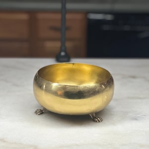 May include: A round, polished brass bowl with a smooth, reflective surface and a wide, open top. The bowl is supported by three small, detailed feet shaped like animal paws, which elevate the base slightly above the surface it rests upon.