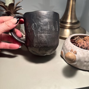 May include: A dark gray ceramic mug with a textured surface and a handle. The mug features an embossed design of stylized trees along the top. The mug is held by a person's hand.