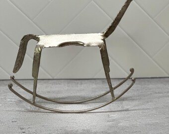 May include: A metallic silver-toned rocking horse figurine crafted from flat metal sheets. The sculpture features a stylized, elongated neck, a simple rectangular body, and a textured tail, all mounted on two curved rockers with curled ends.