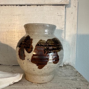 Puede incluir: Un jarrón de cerámica hecho a mano con un cuerpo redondeado y un borde ligeramente ensanchado. El jarrón presenta una base moteada de color blanquecino con patrones marrones oscuros de forma orgánica. El interior está esmaltado y el jarrón se apoya en una base texturizada sin esmaltar. Un objeto decorativo.