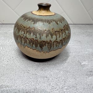 May include: A round ceramic vase with a narrow neck, featuring a beige base color and a decorative drip glaze pattern in shades of muted blue and dark brown that cascades down the upper portion of the rounded body toward the center.