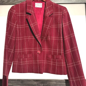 May include: A red plaid blazer with a single-button closure and two front pockets. The blazer is hanging on a wooden hanger. The interior lining is a solid red color. The blazer has a classic, tailored design.