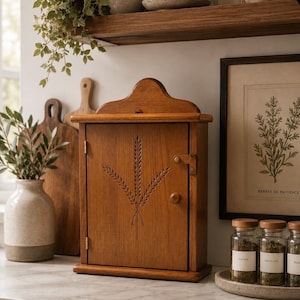 May include: A wooden cabinet with a wheat stalk design on the door. The cabinet is a warm brown color and has a small handle and latch. The cabinet is sitting on a countertop with jars of herbs and a framed botanical print in the background.