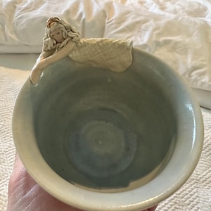 May include: A handmade ceramic bowl with a sleeping angel figurine on the rim. The bowl has a light blue interior and a cream-colored exterior. The angel has long, flowing hair and is wearing a simple gown.