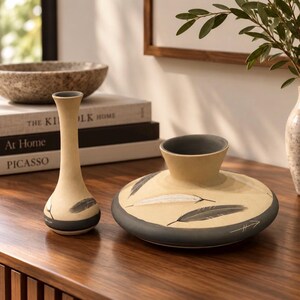 May include: Two ceramic vases with a matte beige finish and dark gray accents. The taller vase has a slender neck, while the shorter, wider vase features a flat, disc-like base. Both pieces are decorated with painted feather motifs and arrow symbols.