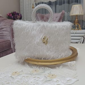 Könnte beinhalten: Eine weiße Kunstfell-Handtasche mit einem abgerundeten Griff, der mit kleinen weißen Perlen besetzt ist. Die Vorderseite der Tasche ist mit einem zentralen, goldfarbenen Metallschmuck in Form eines stilisierten Blattes oder Flügels verziert.