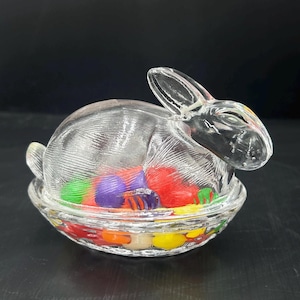 May include: A clear glass bunny-shaped candy dish filled with colorful jelly beans. The bunny has detailed features and sits atop a matching glass dish. The dish is a festive Easter decoration.