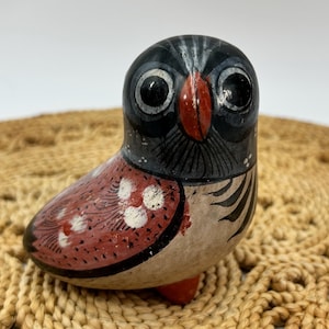 May include: A small, hand-painted ceramic owl figurine featuring a black head with large white-rimmed eyes and a red beak. The body is decorated with a reddish-brown wing pattern, white circular spots, and black feather-like markings on a cream base.