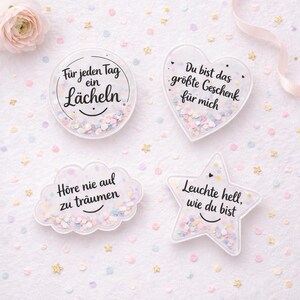 May include: Four clear, decorative coasters with confetti and text. One is round with "Für jeden Tag ein Lächeln", another is heart-shaped with "Du bist das größte Geschenk für mich", a cloud with "Höre nie auf zu träumen", and a star with "Leuchte hell, wie du bist".