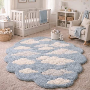 Op de afbeelding: Een lichtblauw, wolkvormig tapijt met witte wolkenaccenten in een kinderkamer. Het tapijt ligt op een neutraal gekleurd tapijt, met een witte wieg, schommelstoel en planken op de achtergrond. De kamer is ingericht met wolken- en sterrenthema's.