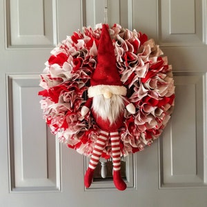 May include: A red and white fabric Christmas wreath with a gnome figure in the center. The gnome is wearing a red hat with a white beard and has red and white striped legs.