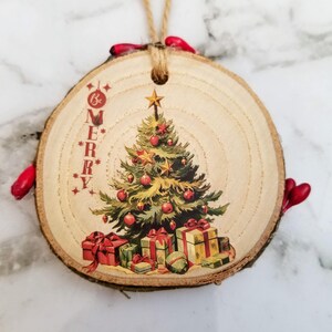 May include: A round wooden Christmas ornament with a printed vintage Christmas tree design. The tree is decorated with red ornaments and gold stars, with wrapped gifts at the base. The text "Be Merry" is printed vertically on the left side. A jute string is attached for hanging.