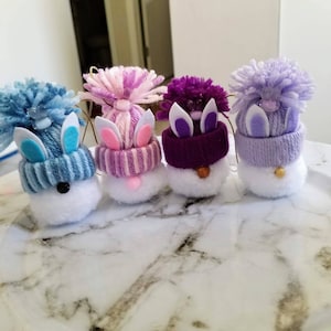 May include: Four handmade snowman ornaments with bunny ears. The snowmen are wearing different colored hats with white pom-poms. The hats are blue, pink, purple, and lavender.