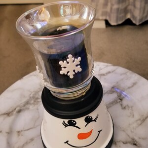 May include: A snowman-themed candle holder. The base is white with a painted face, black hat, and a black rim. A clear glass votive holder with snowflake accents sits atop, holding a dark blue candle.