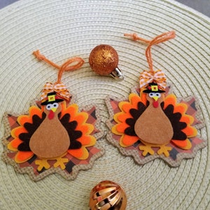 May include: Two hanging ornaments shaped like maple leaves, each featuring a layered felt turkey with a black hat, orange and white gingham bow, and orange twine loop. A small glittery orange sphere and a metallic orange sphere are also present.