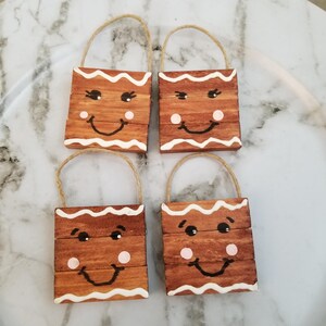 May include: Four wooden gingerbread man ornaments with jute handles. Each ornament has a brown base with a white icing border, black eyes, a pink cheek, and a smiling mouth. The ornaments are arranged on a white surface.