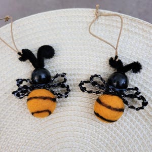 May include: Two handmade bee ornaments. Each bee has a round, orange body with black stripes, a black head, and black and white wings. They are suspended by a natural twine string. The background is a woven, light-colored surface.