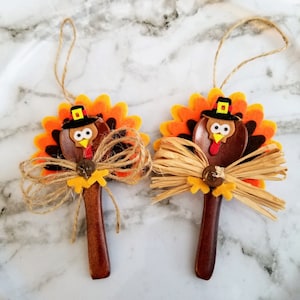 May include: Two wooden spoon turkey ornaments with orange, brown, and yellow felt feathers, a brown felt head, and a yellow felt beak. Each turkey has a brown felt hat with a gold buckle and a brown felt bow tied around the neck.