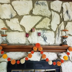 May include: A fireplace mantel decorated with a garland of orange, yellow, and white pom-poms. The garland is strung across the mantel, which is made of wood and has a stone surround. There are three candle holders on the mantel, two with red candles and one with a clear glass candle.