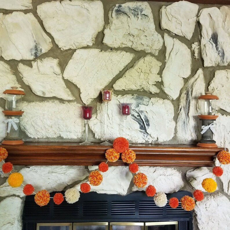 May include: A fireplace mantel decorated with a garland of orange, yellow, and white pom-poms. The garland is strung across the mantel, which is made of wood and has a stone surround. There are three candle holders on the mantel, two with red candles and one with a clear glass candle.