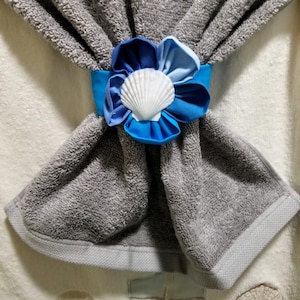 May include: A gray towel folded and held together with a blue ribbon and a decorative flower. The flower has blue petals and a white seashell in the center. The towel is made of a soft, absorbent material.