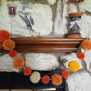 May include: A garland of orange, yellow, and white pom-poms hangs above a fireplace. The garland is made of yarn and is strung together with white thread. The fireplace is made of stone and has a wooden mantel.