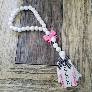 May include: White wooden bead garland with a pink bow and a white tag with black and white checkered ribbon and the text "Shhh!" in black letters.