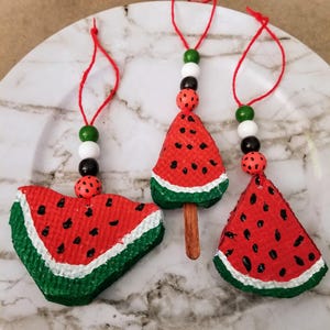 May include: Three handmade watermelon slice ornaments. Each ornament is red with black seeds, a green rind, and white accents. They hang from red string with decorative beads. One is a popsicle shape.