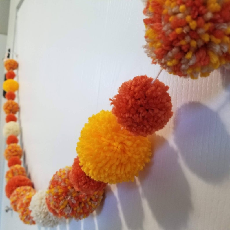 May include: A garland of orange, yellow, and white pom-poms strung together with white thread. The garland is hanging on a white wall.