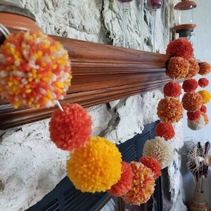 May include: A garland of orange, yellow, and white pom-poms hangs on a wooden mantel. The garland is made of fluffy yarn and is a festive decoration for a home.