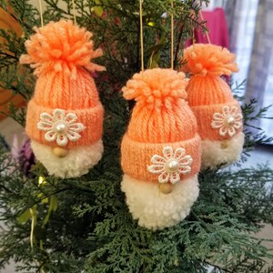 May include: Three peach-colored knitted gnome ornaments with white pom-poms and white lace flowers with pearl centers. The gnomes are hanging from a Christmas tree.
