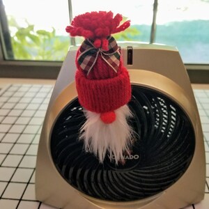May include: A red knitted gnome with a white beard and a plaid bow on its hat, sitting on top of a silver space heater with a black fan.