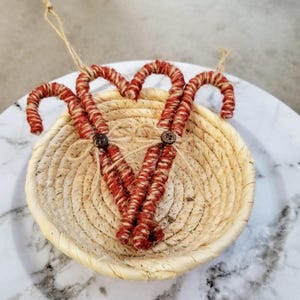 May include: Three handmade candy cane ornaments with red and cream-colored twine wrapped around them. The candy canes are attached to a woven basket with twine and wooden buttons. The ornaments are hanging from twine loops.