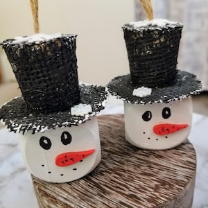 May include: Two white snowman ornaments with black top hats and red noses. The snowman ornaments are decorated with white glitter and black paint.