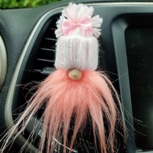 May include: A pink and white gnome with a pink bow on its hat. The gnome is hanging from a car's air vent.