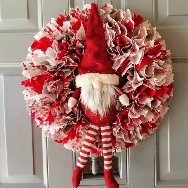 May include: A red and white fabric Christmas wreath with a gnome figure in the center. The gnome is wearing a red hat with a white trim, a white beard, and red and white striped pants.