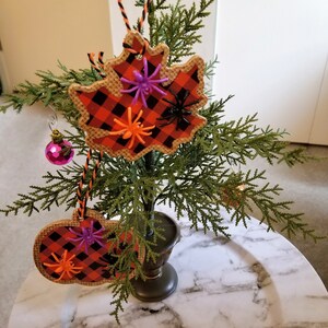 May include: A small, artificial evergreen tree decorated with Halloween-themed ornaments. The ornaments include leaf-shaped and pumpkin-shaped cutouts with a red and black plaid pattern, adorned with plastic spiders in various colors. A pink, faceted ball ornament is also present.