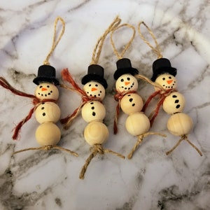 May include: Four wooden bead snowman ornaments with black top hats, orange painted smiles, and red and white yarn scarves. The snowmen are strung with twine for hanging.