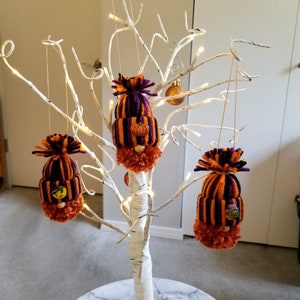 May include: Three Halloween gnome ornaments with orange and purple striped hats and orange beards hang from a white decorative tree. Each gnome has a small pumpkin or Halloween-themed button on its hat. The tree is lit with small white lights.