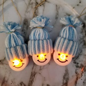 May include: Three white snowman ornaments with light blue and white striped yarn hats. The snowmen have smiling faces and light up with a warm glow.