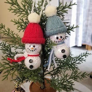 May include: Two snowman ornaments with red and blue knit hats and scarves hang from a small evergreen tree. The snowmen are made of clear spheres filled with white beads, with button eyes and noses. A gold ornament hangs on the tree.