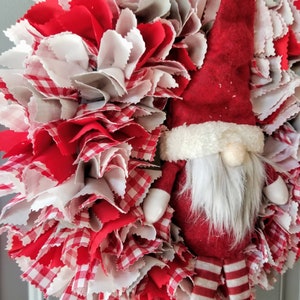 May include: A red and white fabric wreath with a gnome wearing a red hat and white beard. The gnome is holding a white bag in its hand.