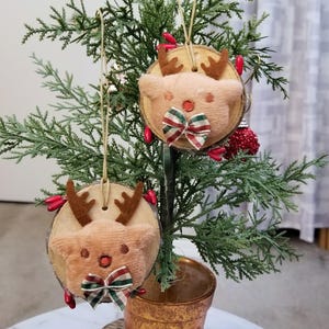 May include: Two Christmas tree ornaments shaped like reindeer. Each ornament is a wood slice with a plush reindeer face, brown antlers, and a plaid bow. The ornaments hang from a small Christmas tree in a gold pot.