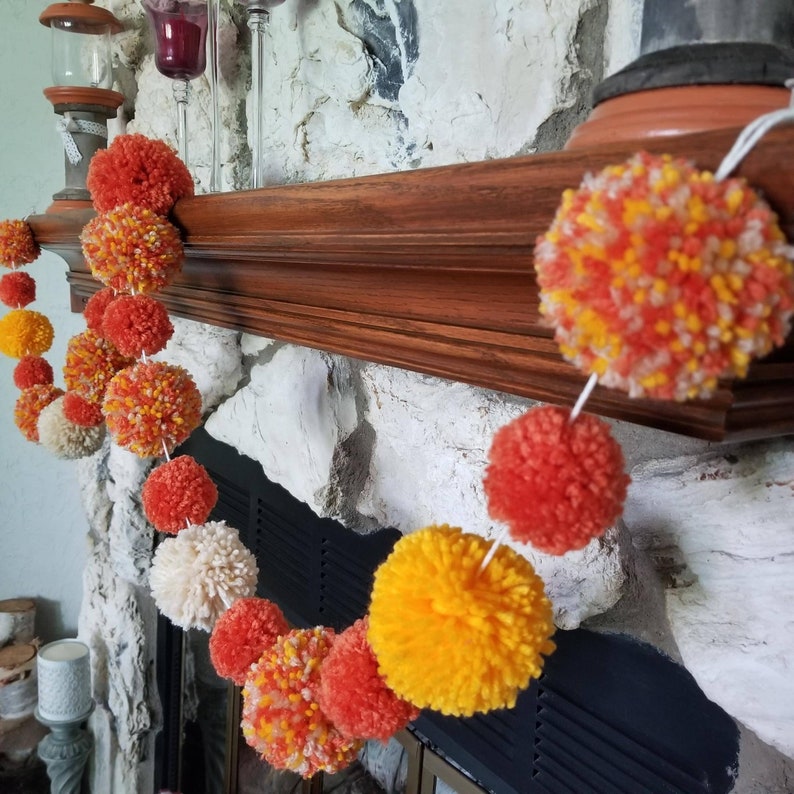 May include: A garland of colorful pom-poms in shades of orange, yellow, and white hangs over a fireplace mantel. The pom-poms are made of yarn and are strung together with white string.