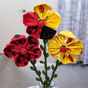 May include: A bouquet of three fabric flowers with ladybug accents. The flowers are red, yellow, and black, with green stems and leaves. Each flower has a ladybug button in the center. The red flower has black polka dots.