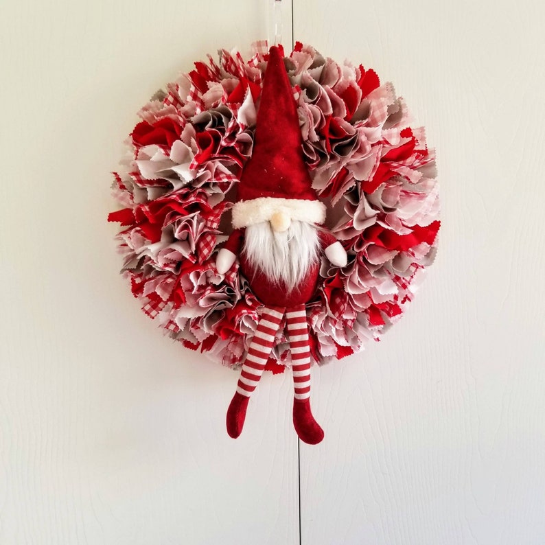 May include: A red and white fabric Christmas wreath with a red gnome with a white beard and red and white striped legs hanging in the center.